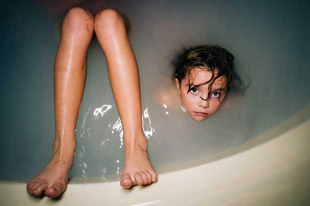 A little girl is showing her face out of a bath tub. There are also a couple of adult legs, the person is out of the camera view